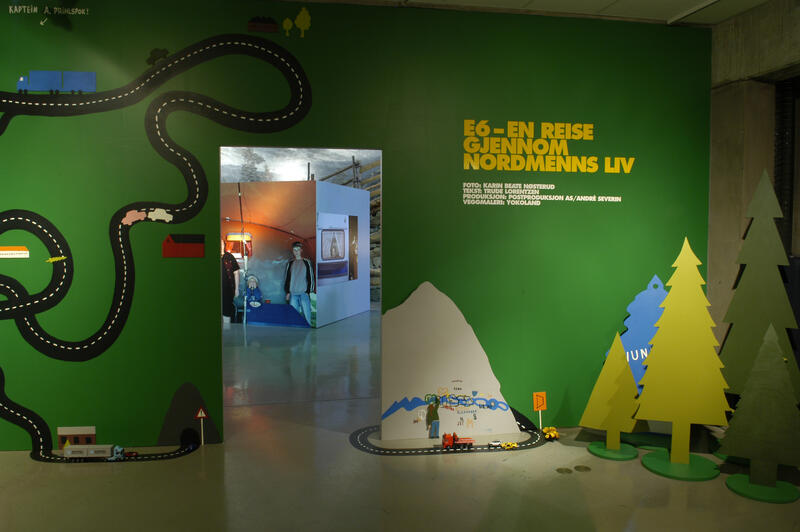 Inngangsparti til utstillingen E6. Grønn vegg med illustrasjon av en vei, noen fjell, samt noen trær (disse står på gulvet foran veggen).