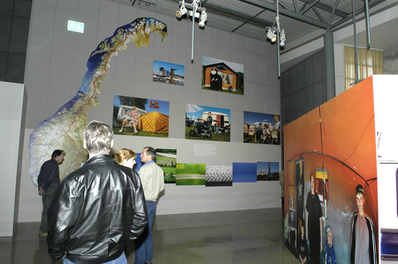 Besøkende ser på utstillingen. På veggen foran de besøkende er det et Norgeskart som viser hele E6-strekningen, samt noen fotografier av campingutstyr.