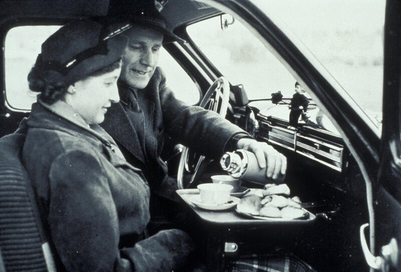 Fotografi av mann og dame med kaffe og småkaker i framsete i bil. Mannen heller varm drikke fra en termokanne opp i en kopp.