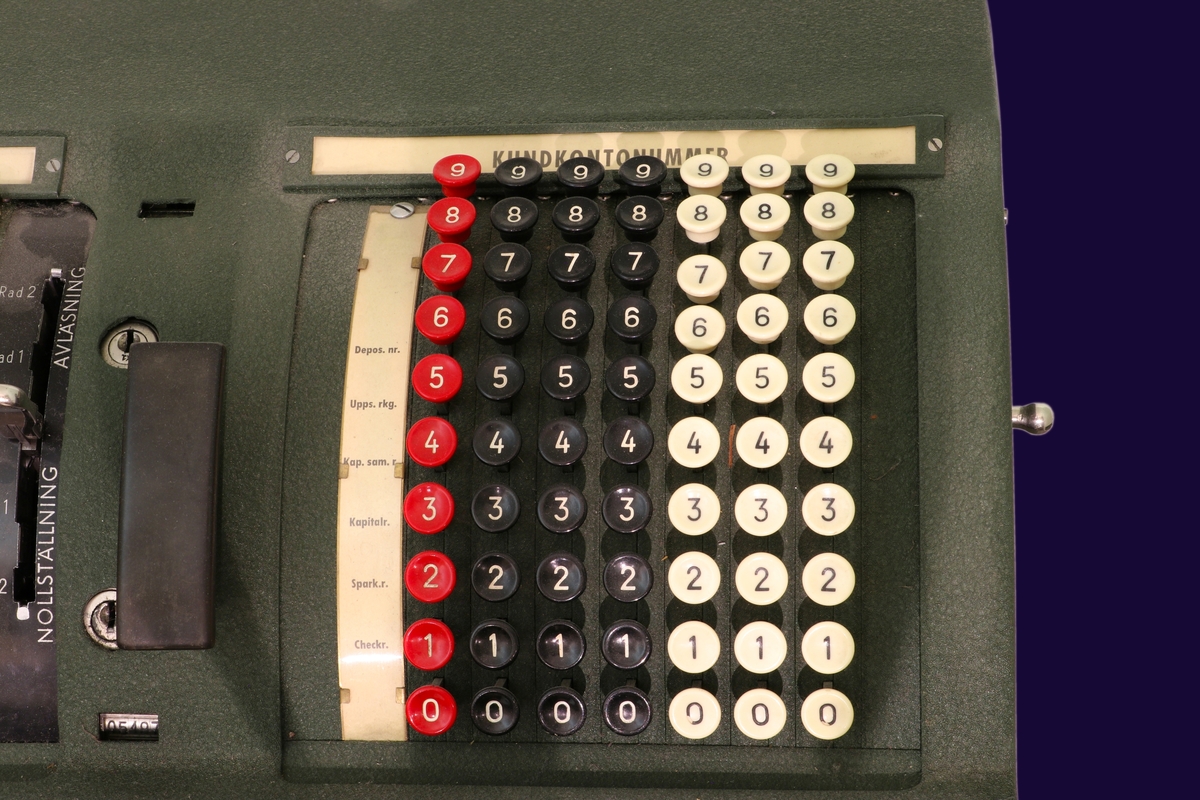 Elektrisk räknemaskin i grön metall. Maskinen står på ett bord med hjul. Siffror och ord på knappar i bakelit i vitt, blått, rött och svart. 
Längst fram på maskinen en tryckdel för att trycka belopp i en bankbok.