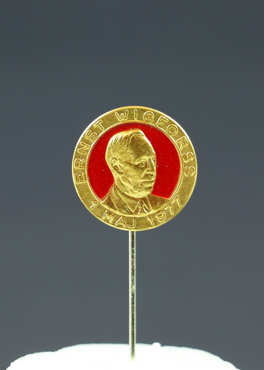 Ett runt nålmärke i guld och rött med präglad text och en man i halvprofil. Längst upp längs med kanten står "Ernst Wigforss" och längs med nedre kanten står det "1 maj 1977".