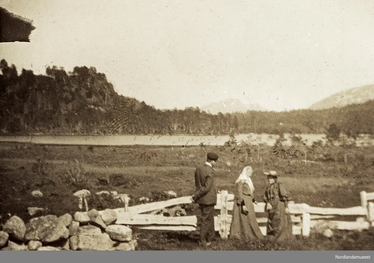 Tre personer ved Ulsetvannet, Tingvoll kommune. De står ved et gjerde av tre og stein. I bakgrunnen skogkledd ås og fjell. Øverst i venstre billedkant ser vi takskjegget på et hus.