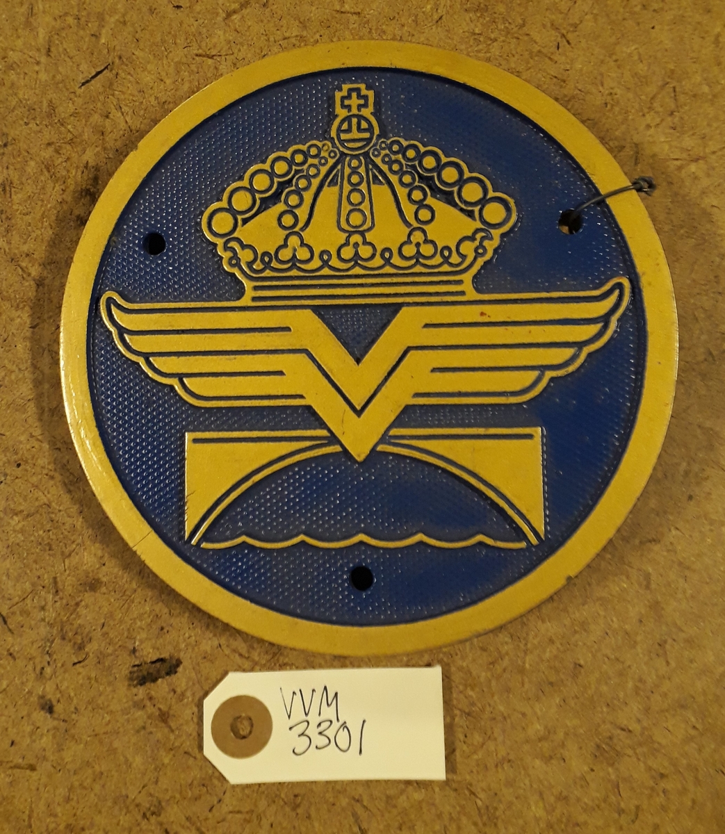 Rund skylt med Vägverkets emblem, av metall med gul kant och dekor på blå räfflad botten. Med tre skruvhål för fastsättning i underlaget.