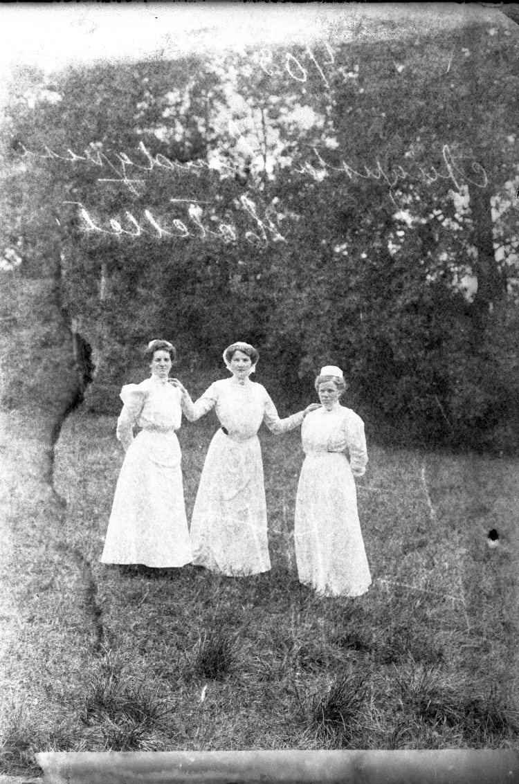 Tre kvinnor i arbetskläder, förkläde och liten mössa på huvudet. Kvinnan i mitten håller sina utsträckta händer på de andras axlar.