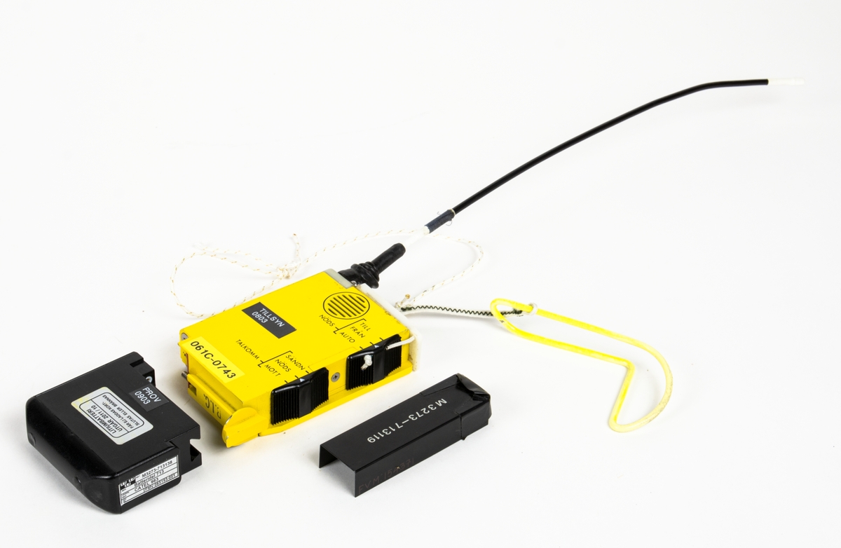 En styck nödsändare med antenn bestående av en sändare, ett batteri, en kåpa till sändaren och en vidhängande krok. Sändaren är i gul metall, rektangulär med 370 mm lång antenn. På ena långsidan finns två avlånga knappar för inställning av nöds: till/från/auto och talkomm: sänd/nöds/mott. Undersidan har en tryckt instruktion av utständning av nödsignal och talkommunikation. Vidhängande gul krok och ett 460 mm långt snöre i plast.

Batteridelen har en påklistrad etikett på kortsidan,
en etikett med text: "PROV 0903" och en påklistrad etikett med text: "LITIUMBATTERI UTGÅR 2011/10".

Kåpa i plast med M-nummer. Påklistrad svart etikett ovansida: "Tillsyn 0803"