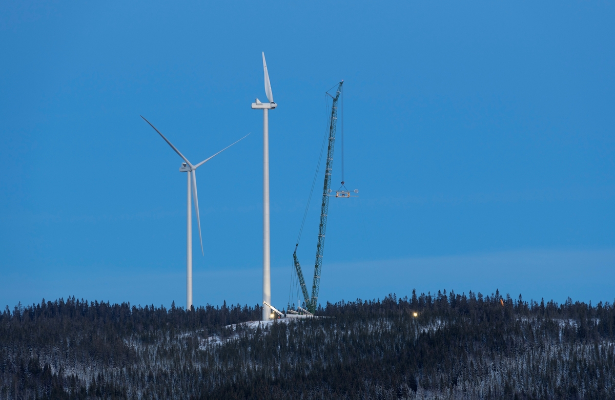Bygging av vindkraftverk på Finnskogen, Hedmark. Kjølberget Vindkraftverk, Våler kommune, Innlandet. Kran heiser et rotorblad under montering av toppdelene på en vindturbin på åsryggen sør for Kjølberget.  Bildet er tatt nær Bjørndamkoia en sen kveld.