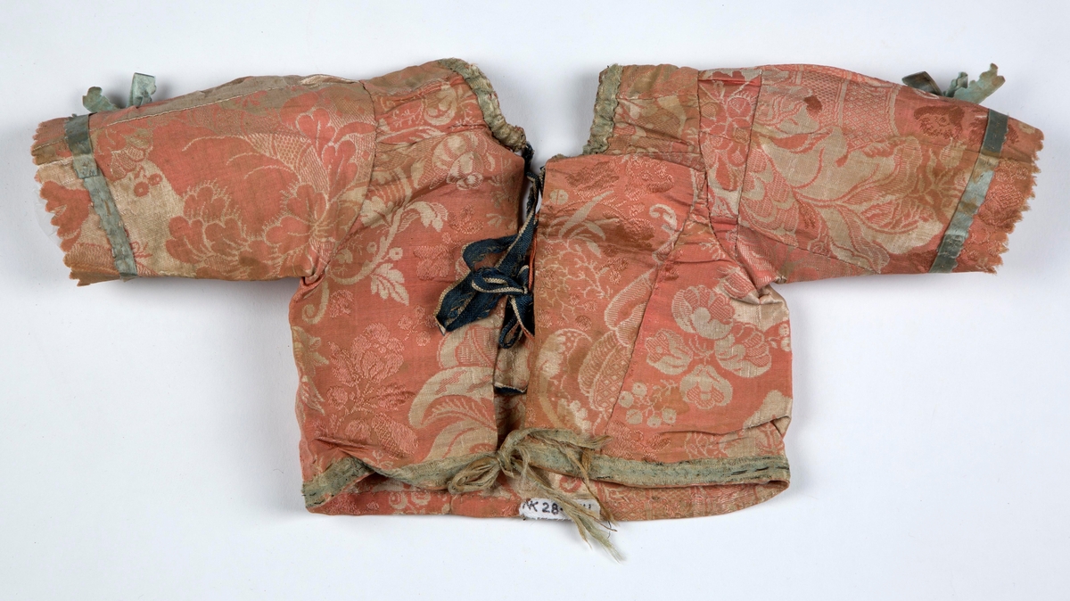Jakken er del av en dåpsdrakt, og sydd av mange små rester av stormønstret kjolestoff fra første halvdel av 1700-tallet. Ermene er relativt rett isatt, og avsluttet med sik-sak-klipping av stoffet. Halsringningen er rund, lavere foran og utstyrt med løpegang. Lukning midt bak med mørkeblå- og hvitstripete lin knyttebånd. Rundt halsringningen og nederst på jakken er påsydd lyseblått silkebånd, som også ender i et knyttebånd bak. Tilsvarende silkebånd, med sik-sak-klippet ytterkant pryder ermene rundt håndleddene, festet med prikkesting og avsluttet med sløyfe på framsiden. Jakken er ikke fôret. Båndene rundt halsen og nederst, er festet med grove tråklesting, og antagelig en senere reparasjon.