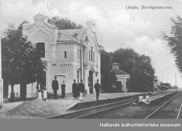 Vykort, "Långås, Järnvägsstationen." Stationen ligger norr om Falkenberg och tillkom längs Mellersta Hallands Järnväg mellan Varberg-Halmstad, invigd 1886. På perrongen står stationspersonal tillsammans med ett par små flickor och mellan spåren sitter tre andra små barn.