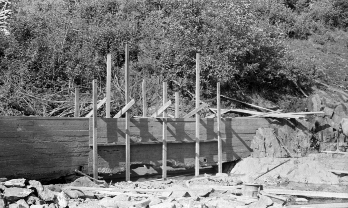 Reparasjon av støpt skådam ved Hartmannsfossen i Haugsåa i Nord-Odal sommeren 1955. Bildet er tatt fra et nesten tørrlagt elveløp på en mur som var oppført ved elvebredden, i et noe berglendt terreng. På skådammen, som åpenbart ikke var ny da bildet ble tatt, var det reist et lite stillas av tre. Det kan virke som om den støpte konstruksjonen var delvis undergravd.