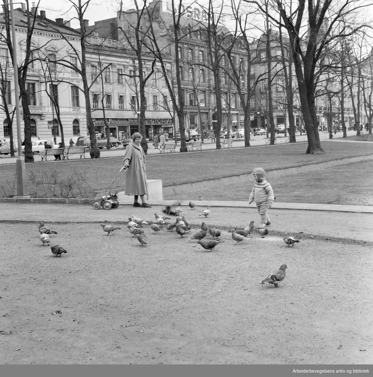 Mor og barn ved Spikersuppa. Fasadene til blant annet Victoria kino i Karl Johans gate 35, Bennetts reisebyrå og Grand Hotel. April 1958.