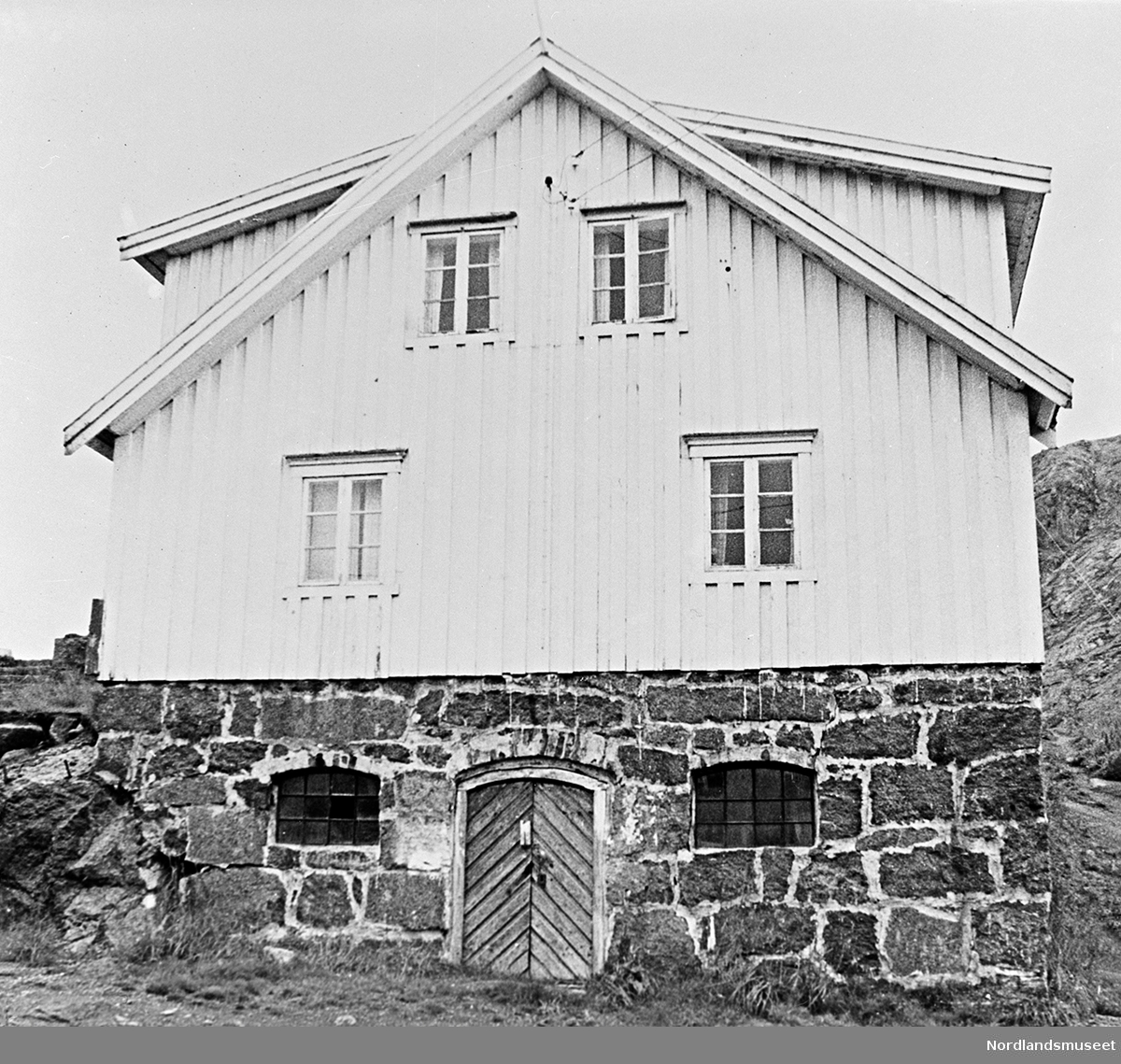 Hvit bolig i fiskeværet Nusfjord. Kortveggen imot. Kjellermur av naturstein med dør og ett vindu på hver side av døra. Smårutede vinduer, stående panel. Deler av taket forsvinner i øvre billedkant.
