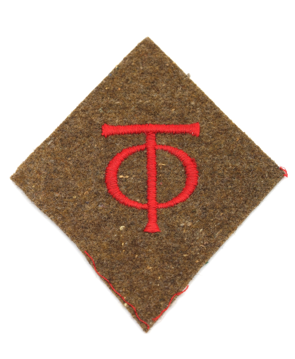 Jakkemerke for organisasjon Todt med initialer brodert/sydd med rød tråd på olivengrønt ullstoff. Til å sy fast til uniform. Ikke nødvendigvis originalgjenstand.