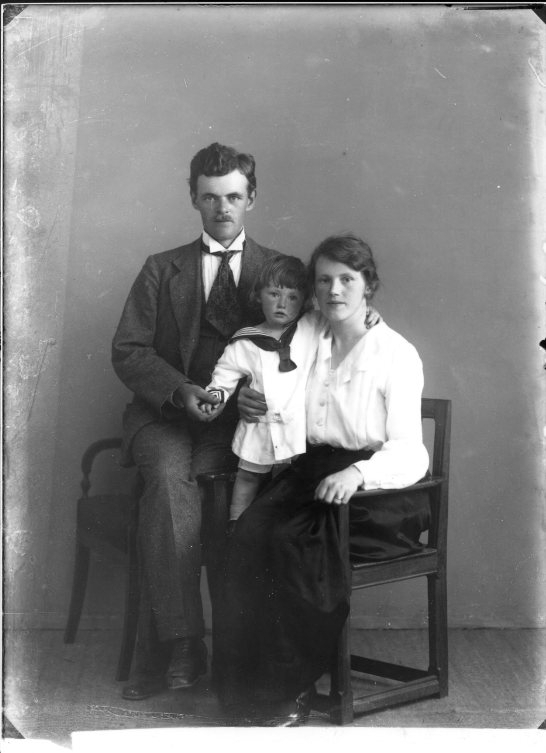 En man sitter på armstödet på en stol tätt intill en sittande kvinna, mellan dem en lite pojke i sjömanskostym.