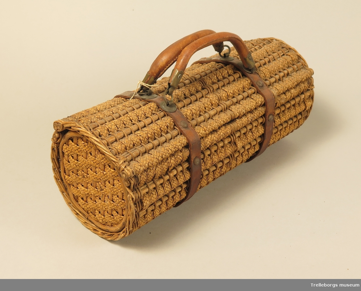 Väska med läderremmar och läderhandtag. Väskans sidostycke utgöres av två st cirkelrunda träplattor. Väskans stomme utgöres av smala pinnar kring vilka det trätts band (0,8 cm) av flätade strån.