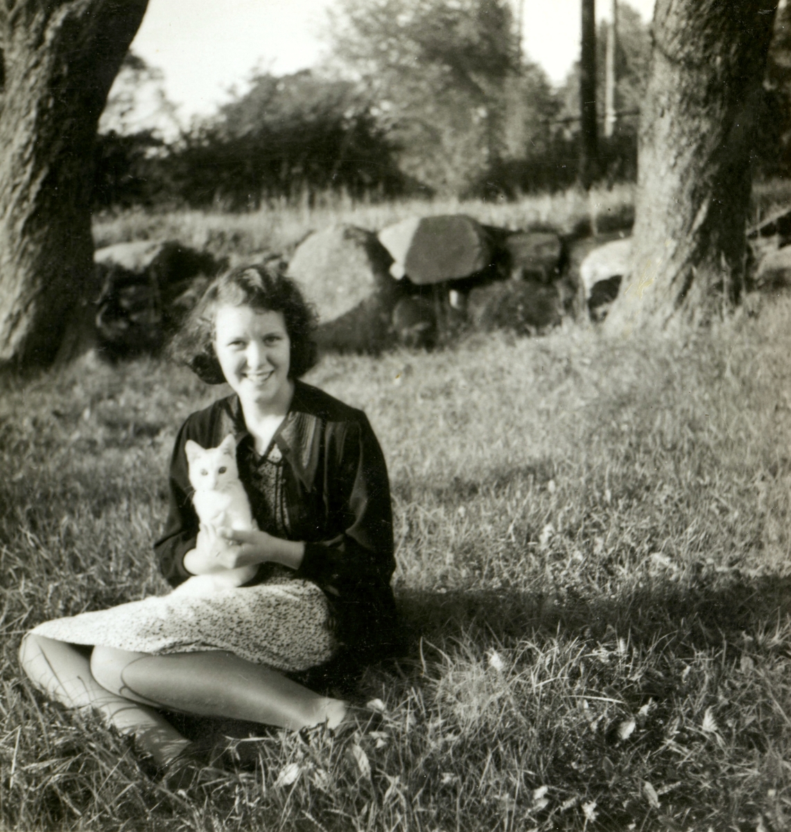 Ingrid Andersson (1918 - 2001, gift Skansing) sitter på gräsmattan hållandes en vit kattunge, Livered 1:15 "Majas" okänt årtal. I bakgrunden ses en gärdesgård.