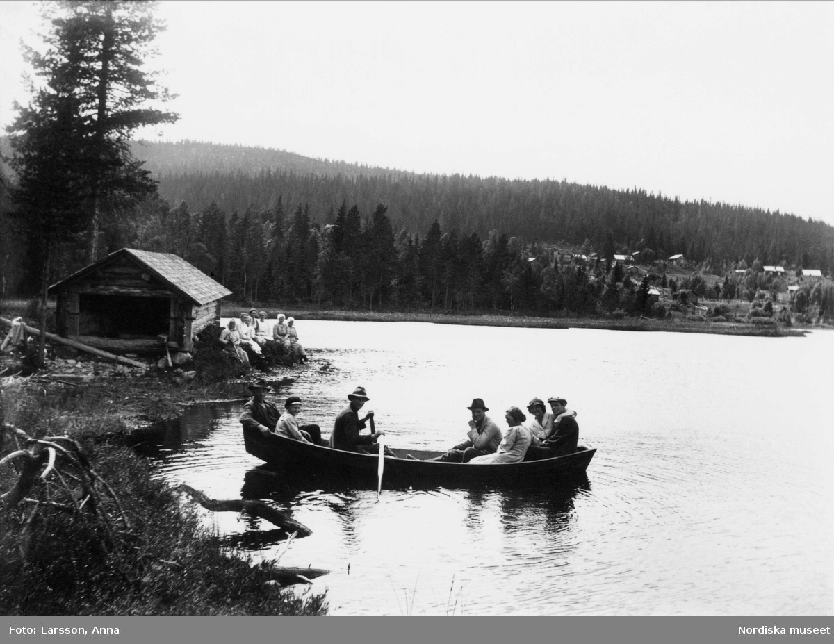 Vid Bårberget, sex personer i båt med berg och båthus i bakgrunden