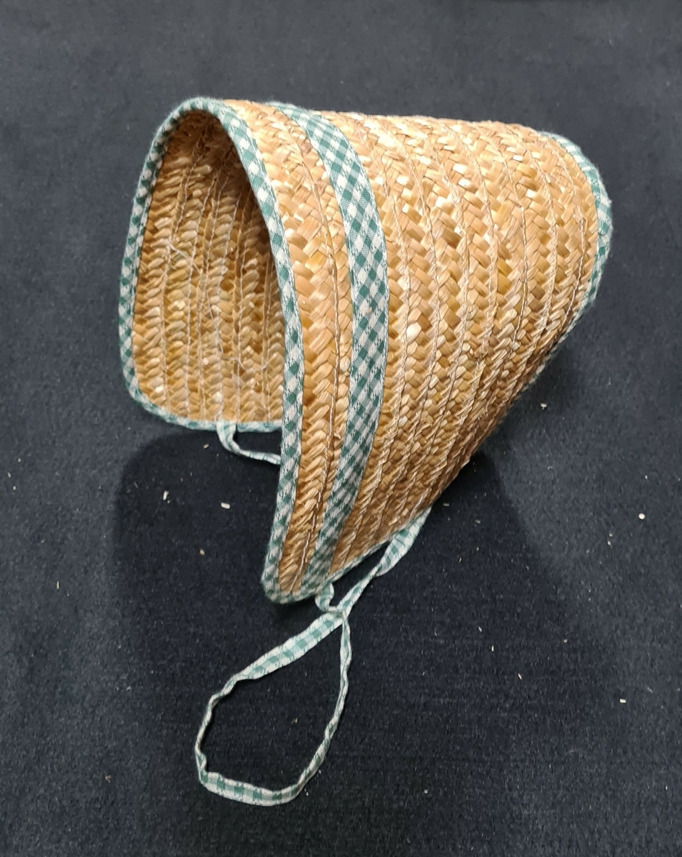 Två halmhattar i formen av en bahytt med hakband. Kantad med grön-vitrutigt tyg. Nacken har samma rutade tyg och även hakbandet. Textilband vid andra raden på bahytten.
