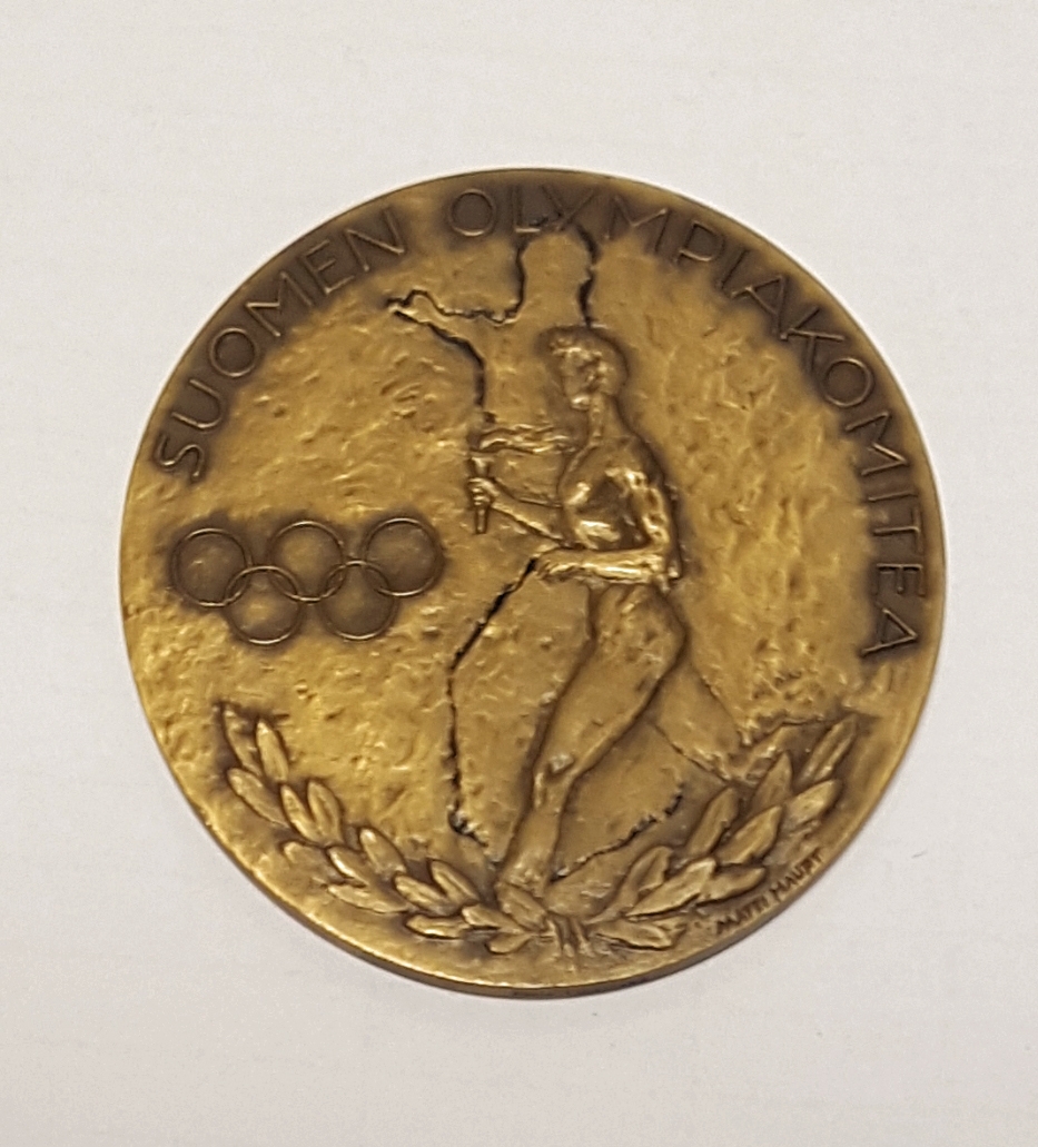 Rund medalje av bronse med en fakkelbærer, de olympiske ringer og olivenkvist. Tekst rundt kanten.