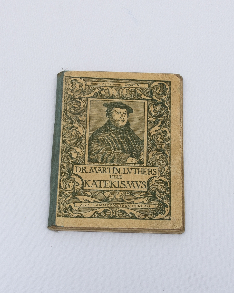 Katekisme "Dr. Martin Luthers lille katekismus" med , et tresnitt av Martin Luther på forsiden, dekorert med bladmønster, innbundet med stive permer og shirtingforsterkelse i ryggen. Sidene og permen er stiftet sammen. 