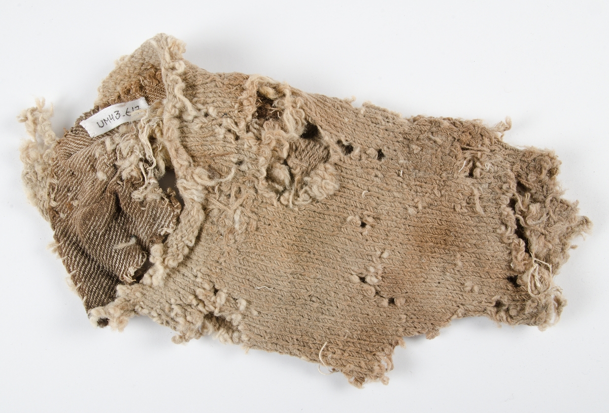 Fragment av socka/strumpa tillverkad av ullgarn. Slätstickning. Många hål, avsaknad av bl.a. tådel och häldel. Lagningar också med fastsydd tygdel, kypertvävd.