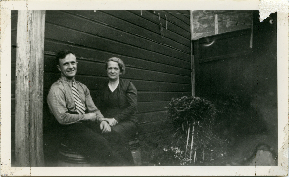 En mann og en kvinne sitter på tønner i solveggen. De ser med glade ansikter mot fotografen. Det hersker en fortrolig stemning. Til høyre i bildet ser man et blomsterbed.