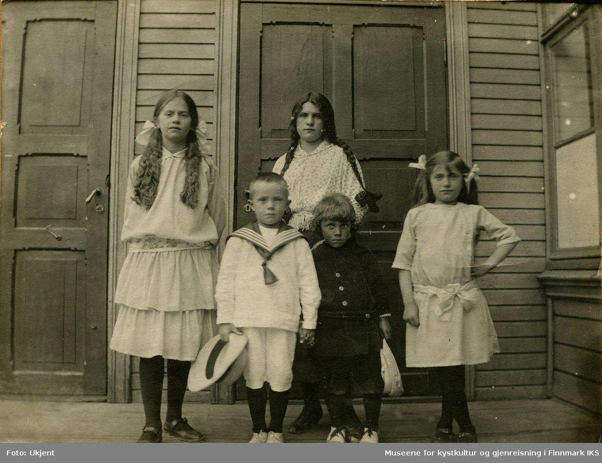 Fem barn står foran en inngangsdør til et trebygg. Fire av dem har lyse finklær på seg mes de har et barn med mørke klær blant seg.