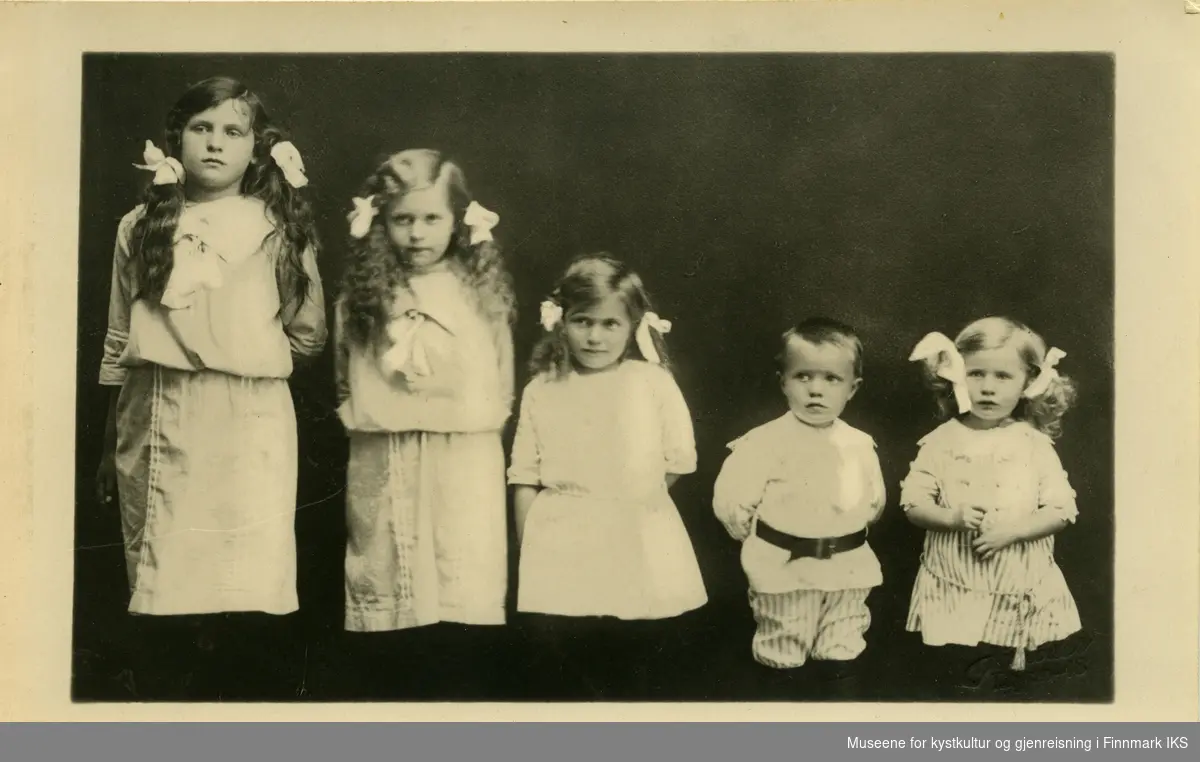 Fem barn har stilt seg opp i sine lyse finklær hos fotografen. Jentene har musefletter med sløyfer.