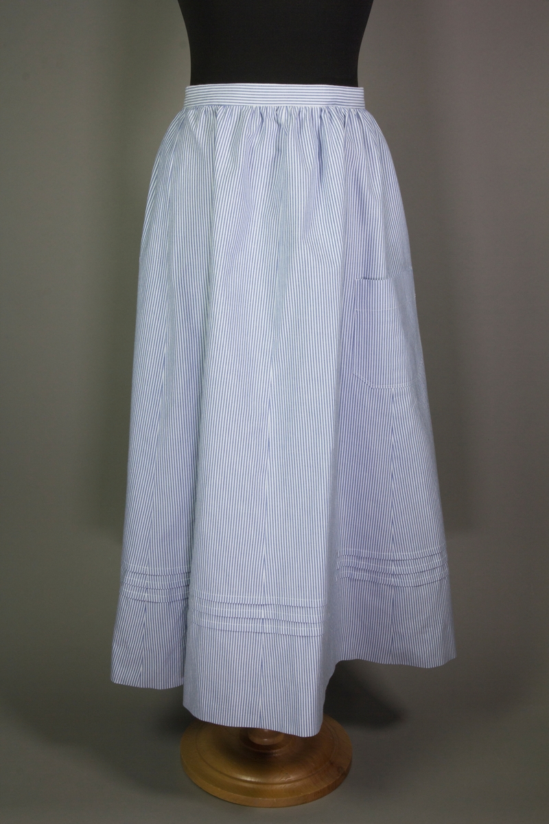 Kjol till tvådelad dräkt av vit och blårandig bomullsväv. Kjolen är hellång med sprund i sidan och tre uppvikta veck.