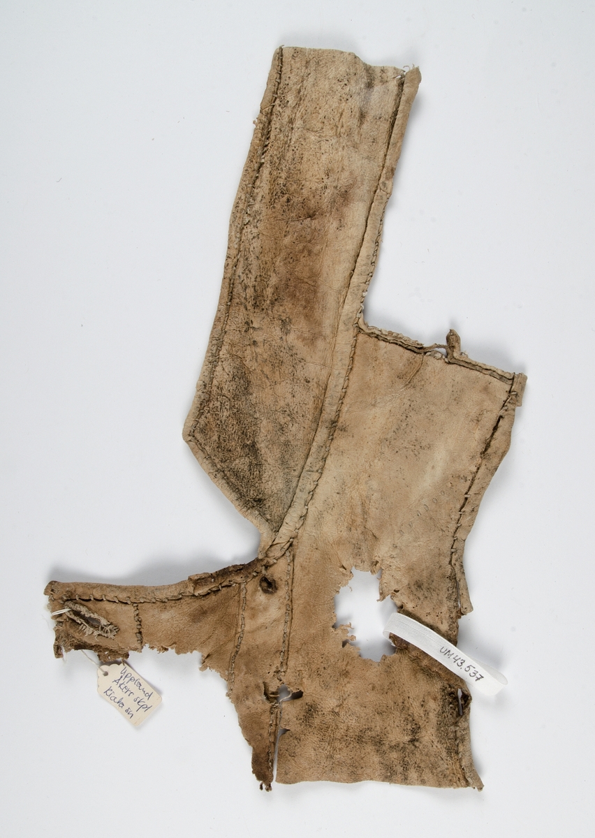 Fragment av pälsplagg, rester av ull på insidan. Sannolikt mansväst med halv ståndkrage och en liten del av framstycket med ärmringing och ett knapphål.