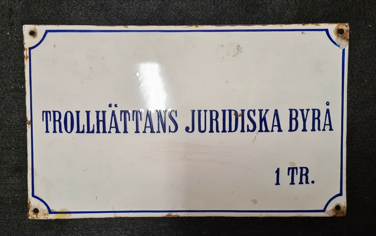 Skylt. Text i blått: Trollhättans juridiska byrå. 1 tr.