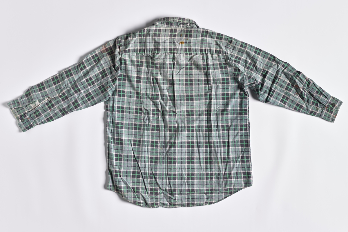 En langermet arbeidsskjorte i bomullsflanell med rutemønster i fargene grønn, grå, hvit og sort. Den har grønne plastknapper. Fabrikkmerke: Triola. Den er fra ca. 1965 og har vært brukt som arbeidsskjorte. Giver foteller at snippene på arbeidskjortene ofte ble slitt. da sprettet man snippene av og snudde på dem. Skjorta fikk da et forlenget liv. Denne skjorte endte som arbeidsskjorte på hytta og snippen er urørt.

Arbeidsklærne er samlet inn i forbindelse med El-samlingen (SBH.EL.G.) Klærne har vært brukt av en av medarbeiderne i El-verket fra 1955 til 2007. Med El-verket menes elektrisitetsforsyningen på Romerike/Akershus.  Fritidsklærne har vært brukt av samme personen.