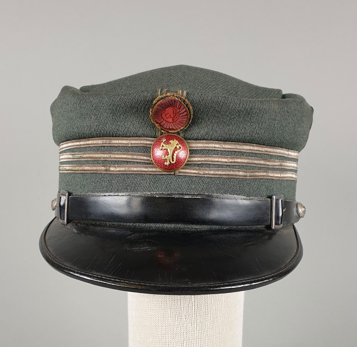 Grønn uniformslue av ull med tre striper rundt pullen, rød knapp med riksvåpen og rosett i front.