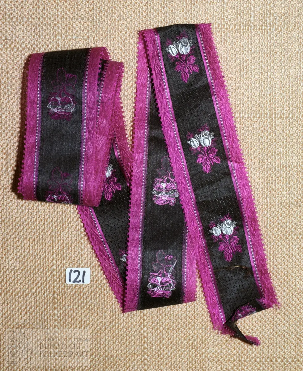 Stripet mønstervevd silkeband i kvitt, rødfiolett og svart med brosjerte blomster i kvitt og rødfiolett langs midtpartiet. Bredde: 5 cm.