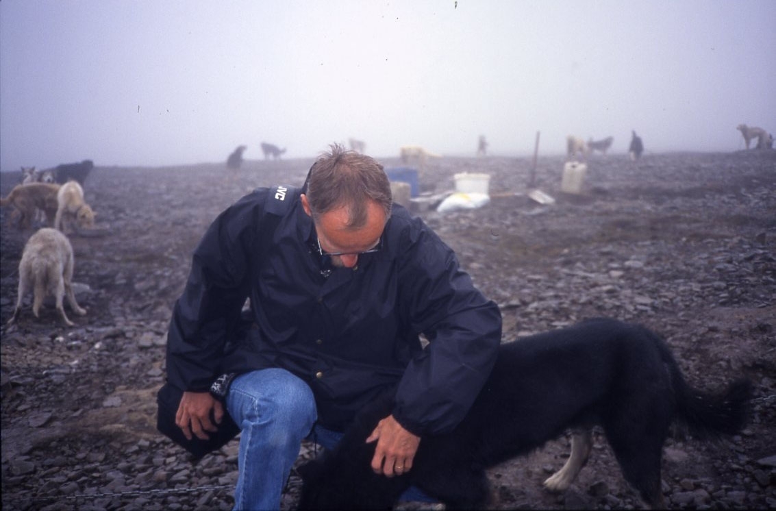 En man sitter på huk och klappar en hund. I bakgrunden ett stort antal hundar på en gård med draghundar, i närheten av Longyearbyen.
