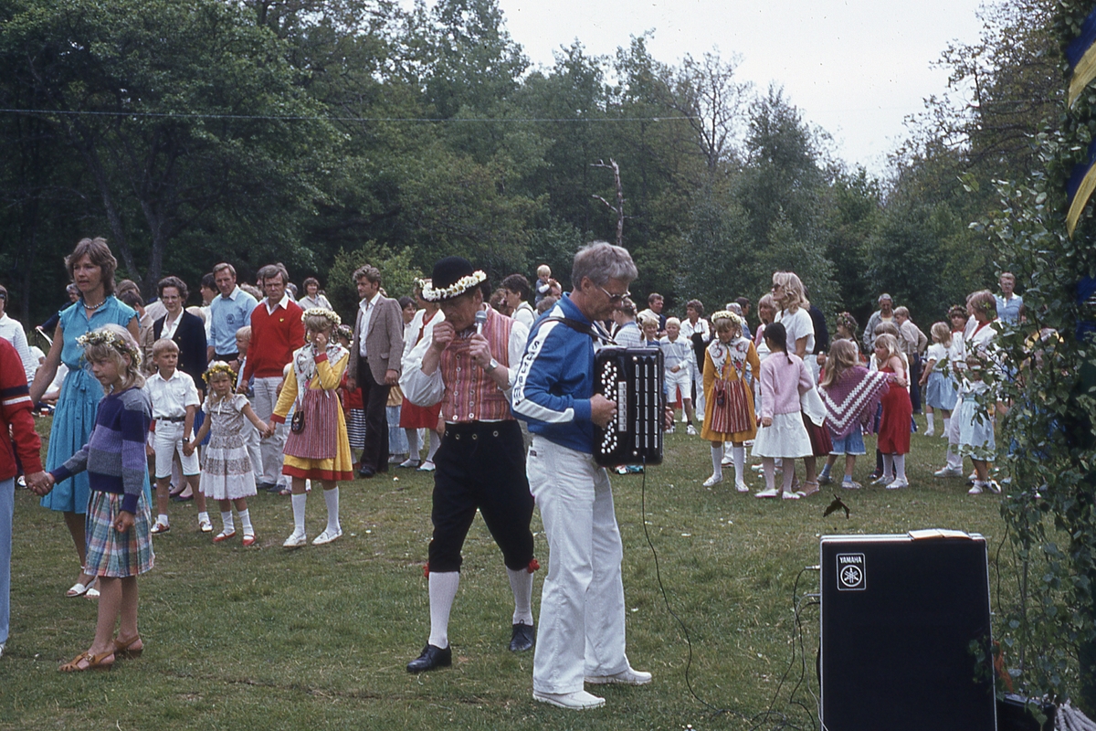 Barn och vuxna dansar kring stången vid midsommarfirande på Ekensås cirka 1982. Sångare/ledare är Sven Olof "Olle" Svensson (klädd i folkdräkt) samt spelmannen Frank Efraimsson som spelar dragspel.
