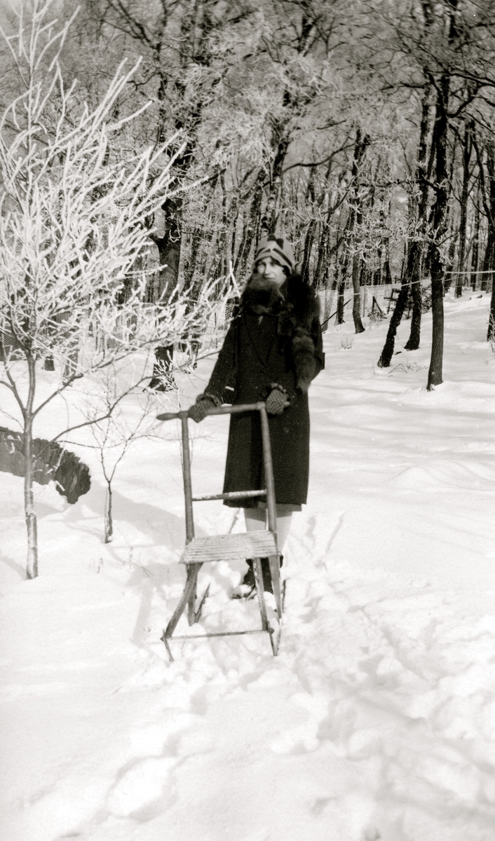 En okänd kvinna står med sin sparkstötting i ett snörikt landskap, Kållered Stom "Nygård" okänt årtal. Till vänster skymtar en hund.