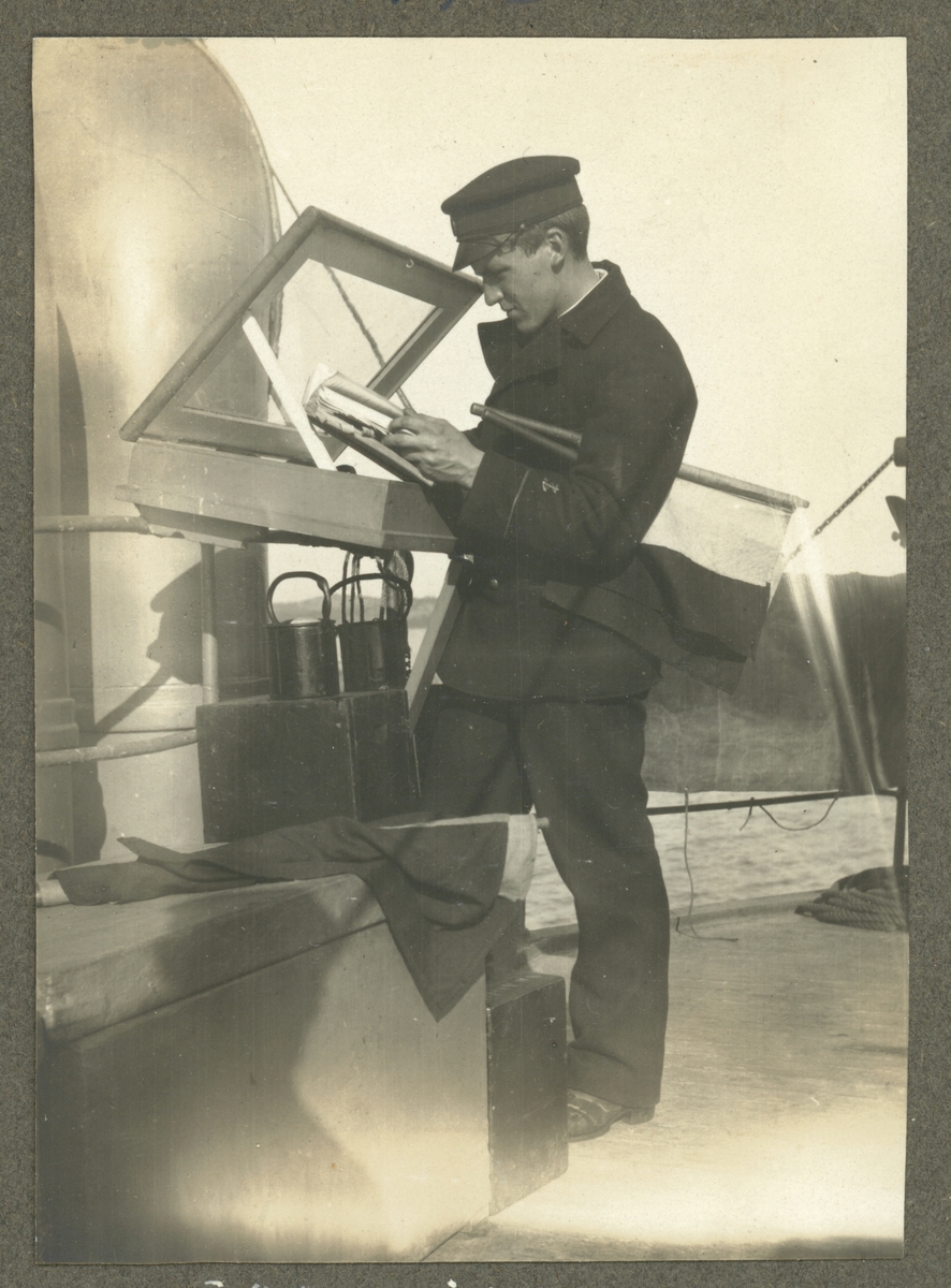 Bilden visar en sjöman på däck av en örlogsfartyg. Han har två signalflaggor under armen och bläddrar i en bok.