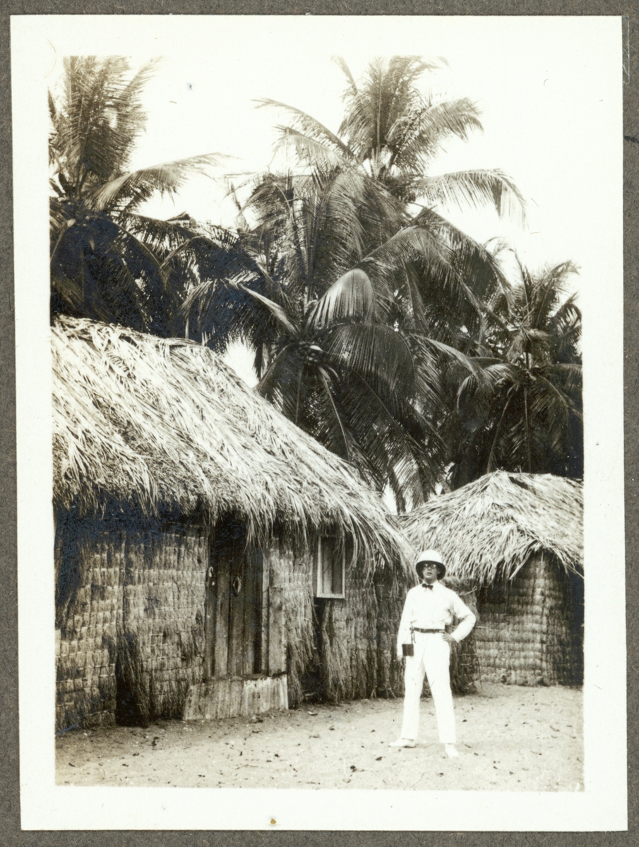 Bilden visar en man med tropikhjälm som står framför an rad hyddor på palmön i Luanda.