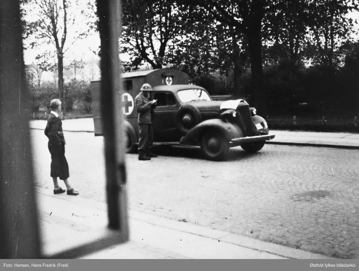 Hjemmestyrker og folkeglede under frigjøringsdagene  i Fredrikstad  1945. Bil på gata og fotgjenger. Røde kors / sanitet. Bilen er en  Chevrolet 1937-modell, ombygd til ambulanse.