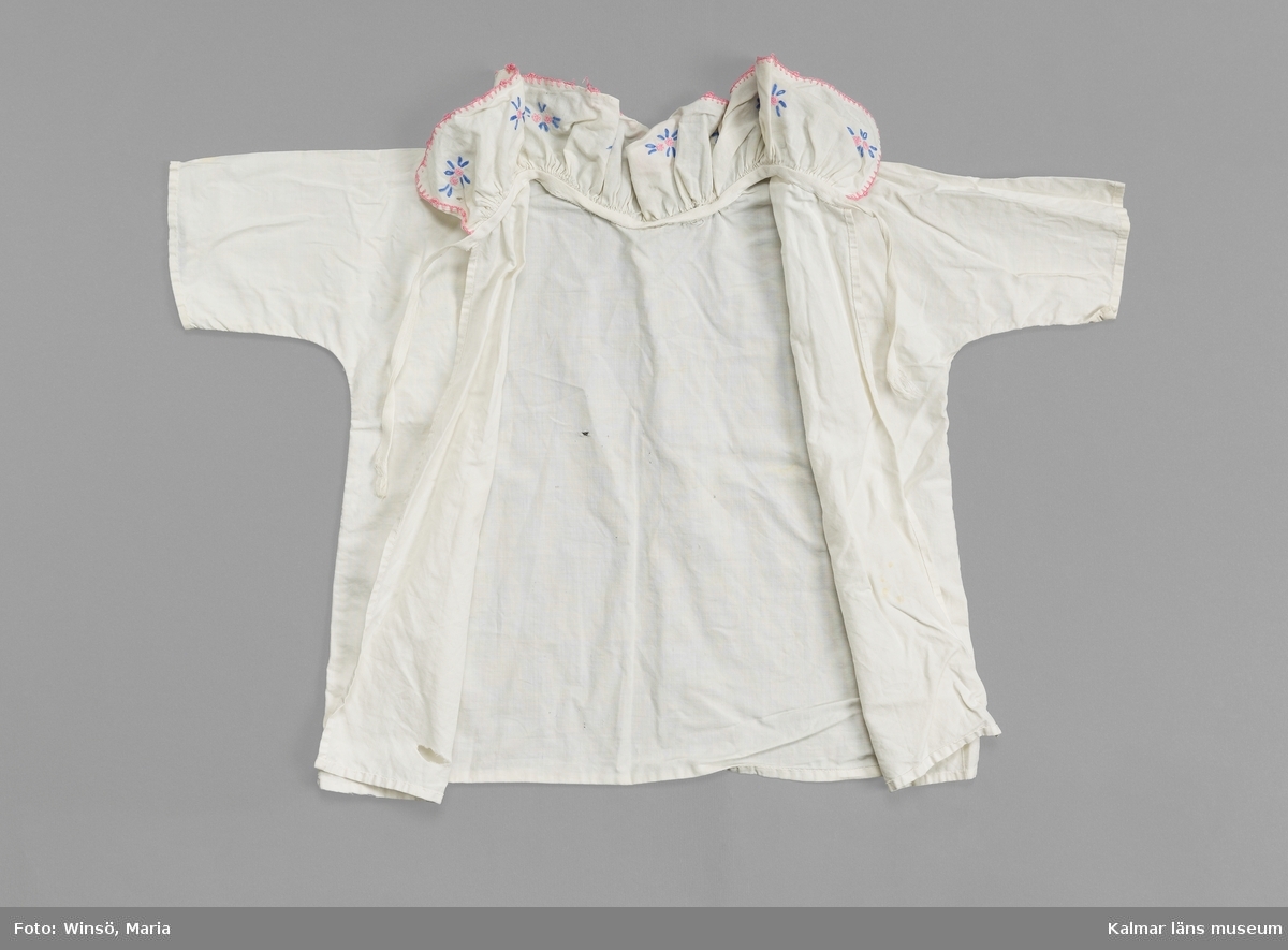 KLM 46489:16. Skjorta, barnskjorta, textil. Barnskjorta av vitt bomullstyg med öppning i ryggen. Knyts med vitt bomullsband i ryggen. Volangkrage med broderade små stjärnliknande mönster i rosa och blått. Broderade stygn runt kragens kant i rosa.