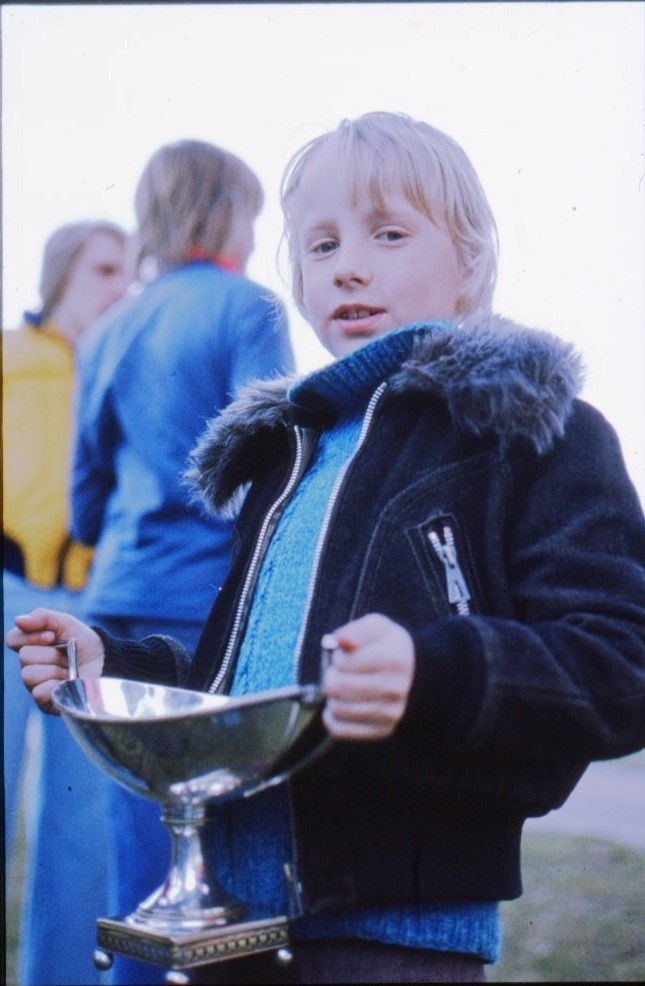 En ljushårig pojke i tjock blå tröja och mörkare blå jacka håller en blå blank bägare i händera, troligen ett ballongpris.