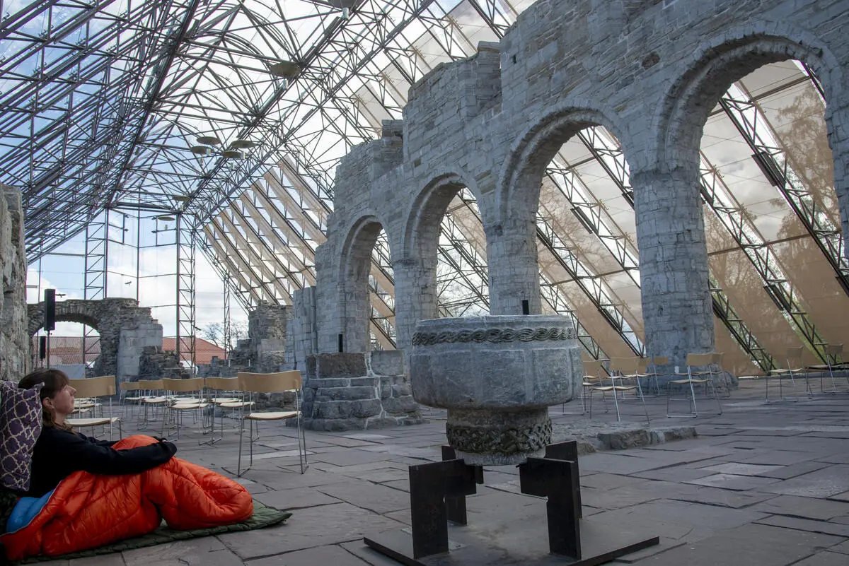 Domkirkeruinen og en middelalderdøpefont beskues av ei kvinne som sitter inni en oransje sovepose med pute bak hodet og lener seg mot den ene steinmuren i Hamardomen.