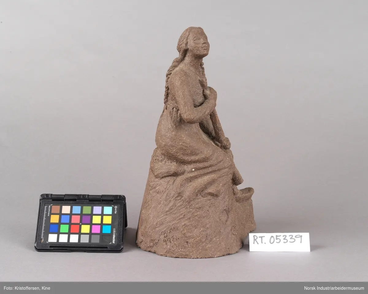 Skulptur laget av leire. Figur, en kvinne med fletter og iført en kjole sitter på en stein med en lur i hendene. Kvinnen vender blikket oppover. Skulpuren er hul.