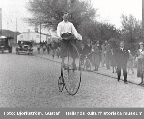 Uppvisning där en man cyklar på en gammal höghjuling, sannolikt på Cykelns Dag i Varberg. Här rullar han förbi åskådrna på Södra vägen vid Rosenfredsskolan. Enligt fotografens liggare är fotot från Sexdagarsloppet det året. Det var en årlig amatörcykeltävling som startades 1924.