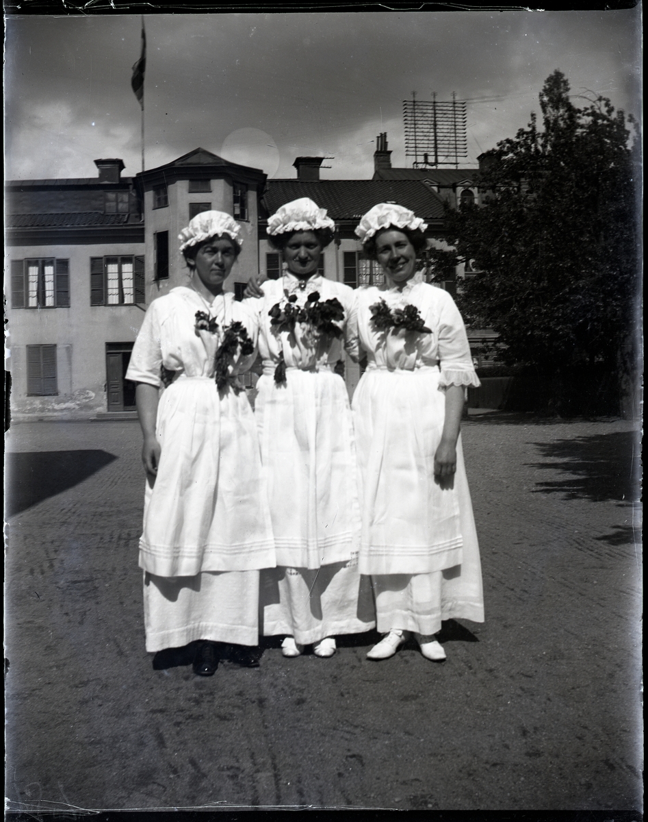 Tre kvinnor med vita husmodersutstyrslar och blommor runt halsen - troligen examen - utanför större byggnad. Gammeldags teleantenner på taket.