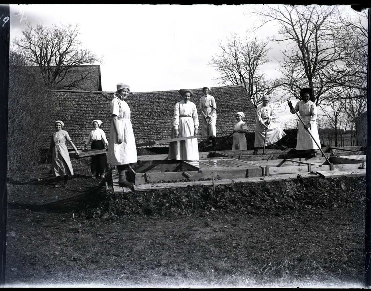 "Husmodersskolan".
Åtta kvinnor arbetar i köksträdgården utanför Husmodersskolan.