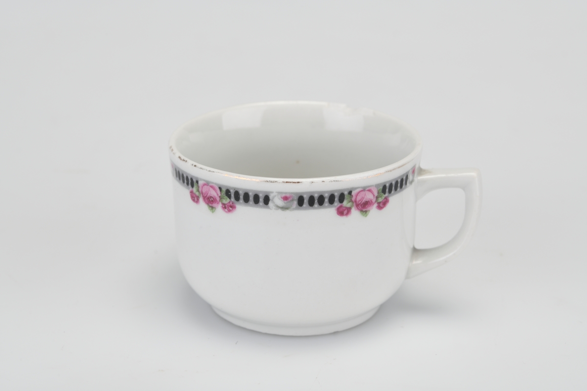 En enkel hvitglasert kopp med en bord øverst. Borden er grå med svarte prikker og små rosa roser med grønne blader. I den ene siden er det en hvit hank.