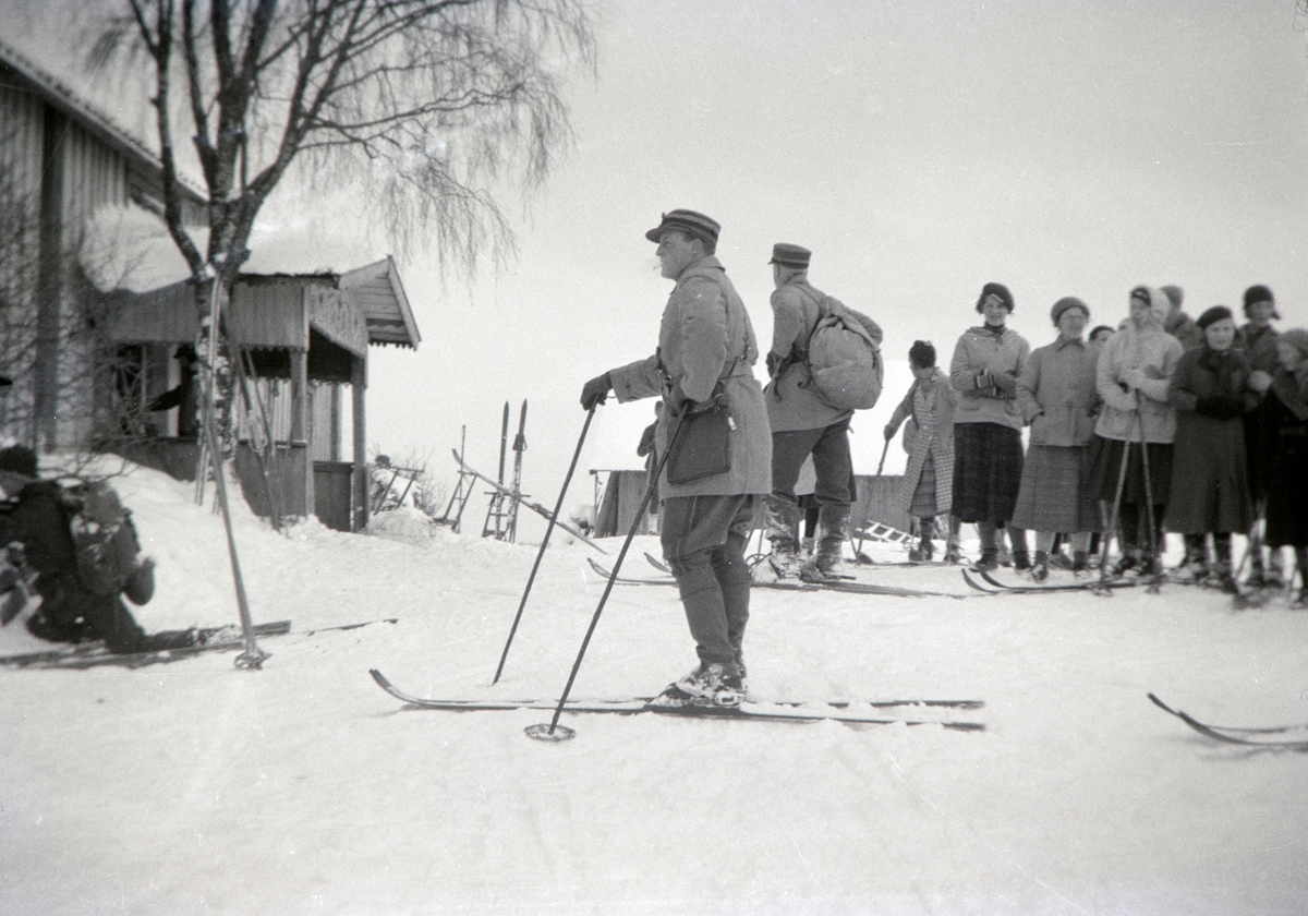 Militærøvelse i Romedal 1937. Vinterøvelse. Kronprins Olav stående på ski foran hovedhuset på Haukåsen. Han var tilstede under øvelsen. Vinter, snø, ski, militæret. Kari Blystad står med mørkt skjørt.