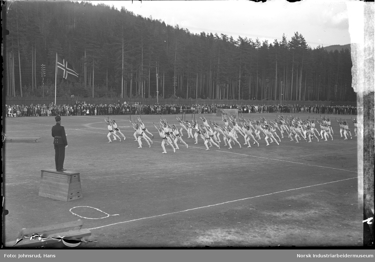 Turnstevne og oppvisning av mannlige turnere på idrettsbane. Mann i uniform stående på kasse på banen. Publikum steående rundt banen.