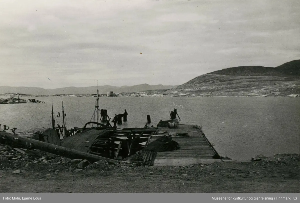 Den ødelagte dampskipskaia i Hammerfest havn etter andre verdenskrig. I bakgrunnen ser man Fuglenes.